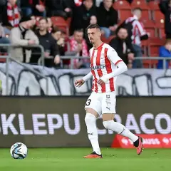 Piłkarz w koszulce z numerem 3 biegnie z piłką. W tle kibice i baner "O SKLEPU CRACOVII!".