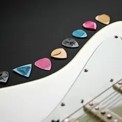Kolorowe kostki do gitary elektrycznej leżą obok białej gitary. Zastanawiasz się, jak nastroić gitarę elektryczną? Zacznij od wyboru odpowiedniej kostki!