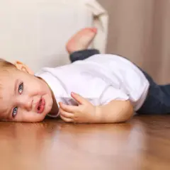 Un bebé de 16 meses con ojos azules y cabello castaño yace en el suelo de madera, vestido con una camiseta blanca y jeans.