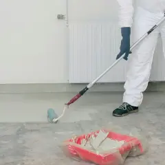 Person i skyddsdräkt målar betonggolv med roller. En färgtråg med ljus färg står på golvet.