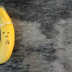 Banan z namalowaną buźką i kciukiem w górę, ubrany w **kondom**. To zabawne przypomnienie o bezpieczeństwie.