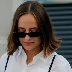 Kobieta z włosami w stylu italian bob, w białej koszuli i czarnych okularach przeciwsłonecznych, patrzy w dół.