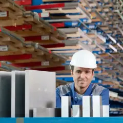 Pracownik w białym kasku stoi przed regałami pełnymi materiałów, obok niego leżą różne stopy aluminium.