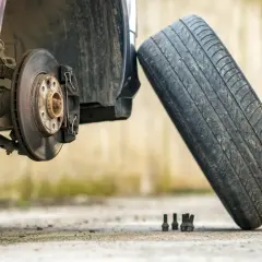 Un coche con el disco de freno expuesto y un neumático viejo apoyado. Se ven tuercas y una llave inglesa en el suelo.