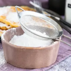 Feiner Puderzucker rieselt aus einem Sieb in eine Schale. So einfach lässt sich puderzucker selber machen für leckere Waffeln.
