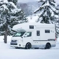 Camping car japonais blanc garé dans la neige, entouré de sapins enneigés. Des remontées mécaniques sont visibles en arrière-plan.