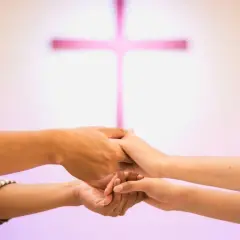 Para młoda trzyma się za ręce, przygotowując się do nauk przedmałżeńskich. W tle różowy krzyż.