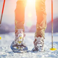 Piernas con raquetas de nieve y bastones listos para excursiones en la nieve bajo un sol brillante.