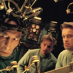 Trzej mężczyźni w dziwnych hełmach, jak z **starych filmów science fiction**, pracują przy skomplikowanej maszynie.