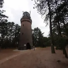 Latarnia morska w Czołpinie, otoczona sosnowym lasem, góruje nad piaszczystym terenem. W oddali widać fragmenty wydm.