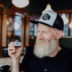 Mann mit Bart hält Tiegel "Beyer's Oil". Perfekt für Bartpflege selber machen.