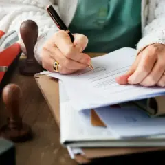 Dłoń pisze na dokumencie, co może być pomocne, jak sprawdzić czy jestem spadkobiercą. Obok stempelki i maszyna do pisania.