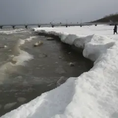 Zimowy krajobraz nad morzem, gdzie woda częściowo zamarza, tworząc lodowe formacje na brzegu. W oddali widać molo i spacerujących ludzi.