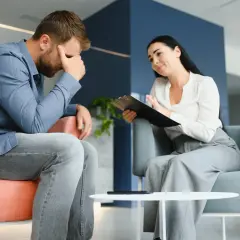 Mężczyzna w niebieskiej koszuli siedzi ze spuszczoną głową, podczas gdy kobieta z notatnikiem mówi do niego.