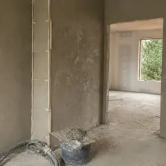 Świeżo nałożony tynk na ścianę w nowym budownictwie. Widać wiadro z zaprawą i kielnię, gotowe do dalszych prac.