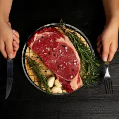 Ojo de bife crudo, sazonado con sal y pimienta, con romero y ajos, listo para cocinar. Manos sostienen cuchillo y tenedor.