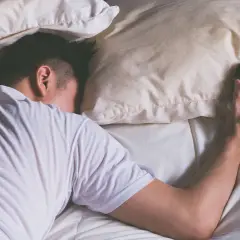 Mann schläft im Bett, Wecker liegt daneben. Wie viel Schlaf braucht man wirklich?