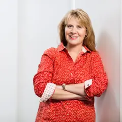 Petra Weber, eine lächelnde Frau mit blonden Haaren, trägt ein rotes Hemd mit Blumenmuster und hat die Arme verschränkt.