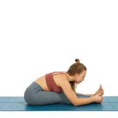 Mujer en esterilla azul, practicando una de las variantes de uttanasana, con las piernas estiradas y el torso inclinado hacia adelante, sujetando los pies.