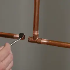 A hand applies flux with a brush to a copper pipe end, preparing it for soldering to a copper tee fitting.