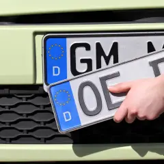 Dłoń trzyma tablice rejestracyjne państw. Jedna z nich ma oznaczenie "GM", druga "OE".