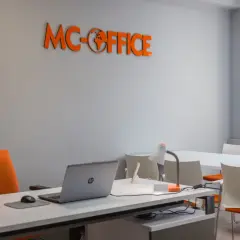 Pomarańczowe logo MC-OFFICE na szarej ścianie nad biurkiem z laptopem.