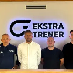 Czterech mężczyzn stoi przed podświetlanym logo "EKSTRA TRENER". Dwóch z nich to srodkowy pomocnik, który jest trenerem.