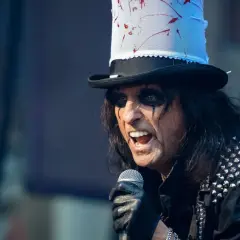 Alice Cooper w Polsce, w swoim charakterystycznym makijażu i z kapeluszem, śpiewa do mikrofonu.