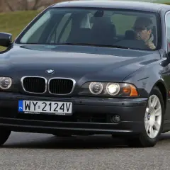 Ciemnoszary sedan BMW E39 z kierowcą w okularach jedzie po drodze.