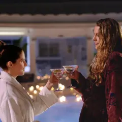 Zwei Frauen stoßen mit Martinis an, umgeben von Kerzenlicht und einem Pool. Nur noch ein kleiner gefallen, bevor der Abend perfekt ist.