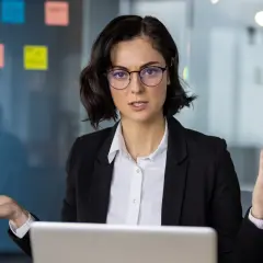 Kobieta w okularach, ubrana w czarną marynarkę, gestykuluje, tłumacząc coś przy laptopie. Na szklanej ścianie w tle widać kolorowe karteczki, jakby z pomysłami na ip box.