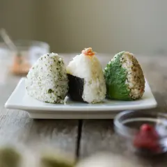 Three onigiri, made of rice, sit on a white plate. One is wrapped in nori, another topped with salmon, and the third with a green leaf.