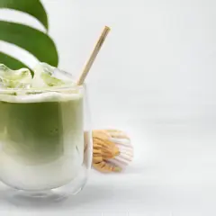 Un verre de matcha à jeun, glacé et crémeux, avec une paille en bois. À côté, un fouet à matcha et une feuille de Monstera.