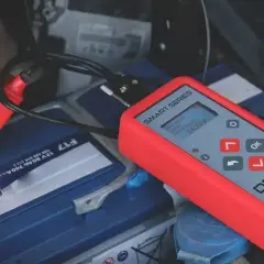Czerwony tester akumulatorów podłączony do akumulatora samochodowego. Wyświetla 14.25V, sugerując, że problemem nie jest rozładowany akumulator, a coś innego.