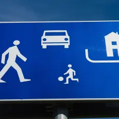 Znak informuje o strefie zamieszkania, gdzie obowiązuje ograniczona prędkość. Widoczni piesi, bawiące się dziecko i samochód.