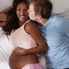 Coppia abbracciata a letto. Lui bacia la guancia di lei, incinta, che sorride. Potrebbe essere un momento di intimità che porta a contrazioni dopo rapporto in gravidanza.