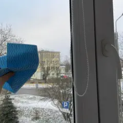 Ręka trzyma niebieską ściereczkę, gotową do mycia okna. Za szybą zimowy krajobraz z drzewami i budynkiem. Czy można myć okna na mrozie?