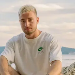 Edzio rap, z blond włosami i w białej koszulce, siedzi na plaży.