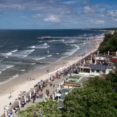 Tętniąca życiem promenada nad morzem, z plażą pełną ludzi, kawiarniami i molo w oddali.