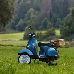 Niebieska Vespa z wiklinowym koszem na bagażniku, zaparkowana na trawie. Wygląda jakby czekała na przegląd skutera po terminie, gotowa na letnią przygodę.