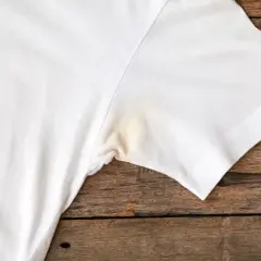 Verkrustete Deoflecken entfernen: Ein weißes T-Shirt mit gelblichen Flecken unter dem Arm auf Holzuntergrund.