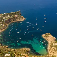 Una baia di Ibiza con numerose barche in fila, tra cui yacht e velieri, su acque cristalline.