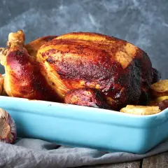Un succulento pollo al forno dorato e croccante, servito in una teglia azzurra con aglio arrosto e patate.