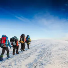 En grupp vandrare med ryggsäckar går uppför en snöig bergssluttning under en klarblå himmel. De njuter av att vandra på vintern.