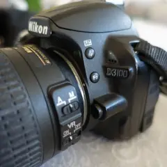 Zbliżenie na aparat Nikon D3100. Widoczny obiektyw z pierścieniem ostrości i przełącznikami AF/M oraz VR ON/OFF. D3100 to model, którego nikon d3100 dane techniczne warto poznać.