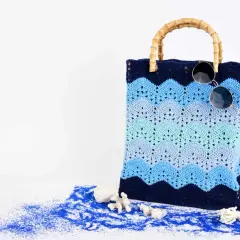 Borsa da spiaggia con manici in bambù, realizzata con splendidi lavori uncinetto cotone a onde blu. Occhiali da sole appoggiati sopra.