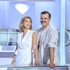 Uśmiechnięci prowadzący koło fortuny, kobieta w białej sukience i mężczyzna w jasnej marynarce, stoją przy stole w studiu.