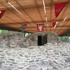 Kamienne ruiny na Ostrowiu Lednickim, z historycznymi chorągwiami i krzyżem. Wyobraź sobie, że to miejsce, skąd wyruszał dawny ostrów lednicki prom.