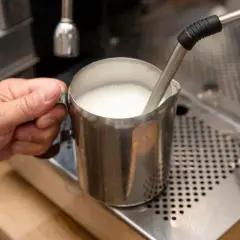 Ręka trzyma metalowy dzbanek z mlekiem, które jest spieniane przez dyszę ekspresu. Tak przygotowane mleko jest kluczowe, czy do cappuccino dodaje się mleko.