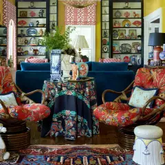 A maximalist living room bursts with color and pattern. Two rattan chairs with vibrant red floral upholstery face a navy sofa. Shelves overflow with decorative plates and vases.